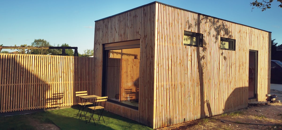 A modern wooden studio is showcased, featuring vertical slats that create a natural aesthetic. Large windows allow for ample natural light, while a small outdoor seating area with a table and chairs invites relaxation. A neatly trimmed lawn surrounds the studio.