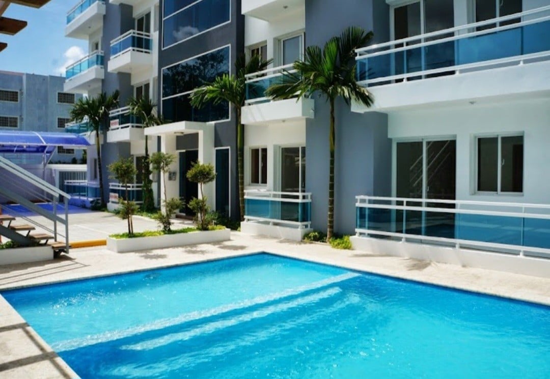 A clear blue swimming pool is situated at the forefront, surrounded by a well-maintained patio. The modern building features multiple balconies and large windows, framed by neatly trimmed palm trees and lush greenery. Sunlight reflects off the water, creating a serene atmosphere.