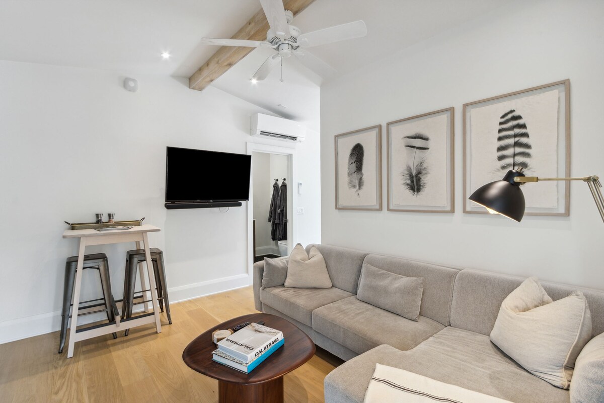 A bright living area features tall ceilings with exposed beams. A comfortable sofa invites relaxation, while a mounted TV offers entertainment. Minimalist artwork adorns the walls, complemented by a wooden coffee table and sleek bar stools near a small dining area.