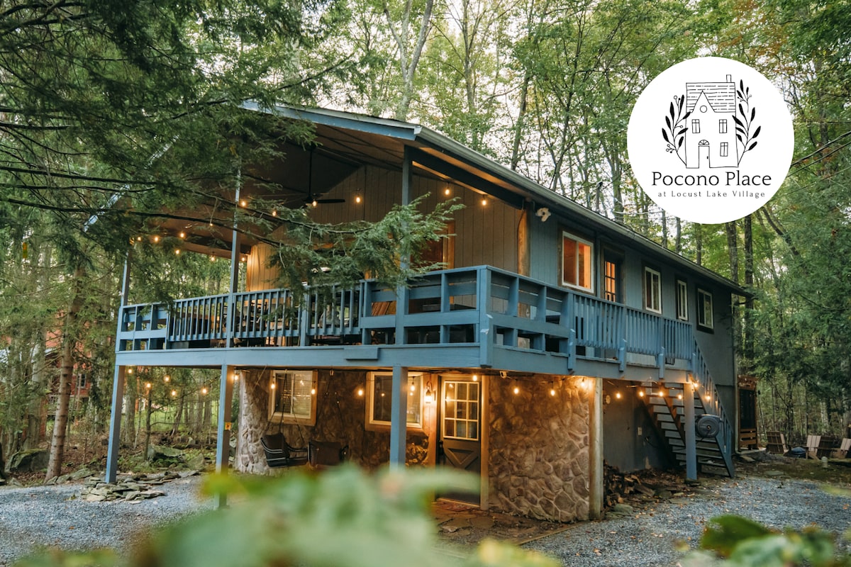 The exterior of the home is shown, featuring a deck above a stone foundation and surrounded by trees. Soft outdoor lighting illuminates the deck, enhancing the inviting presence of the cabin. The entrance is accessible via stairs, leading to a welcoming porch area.