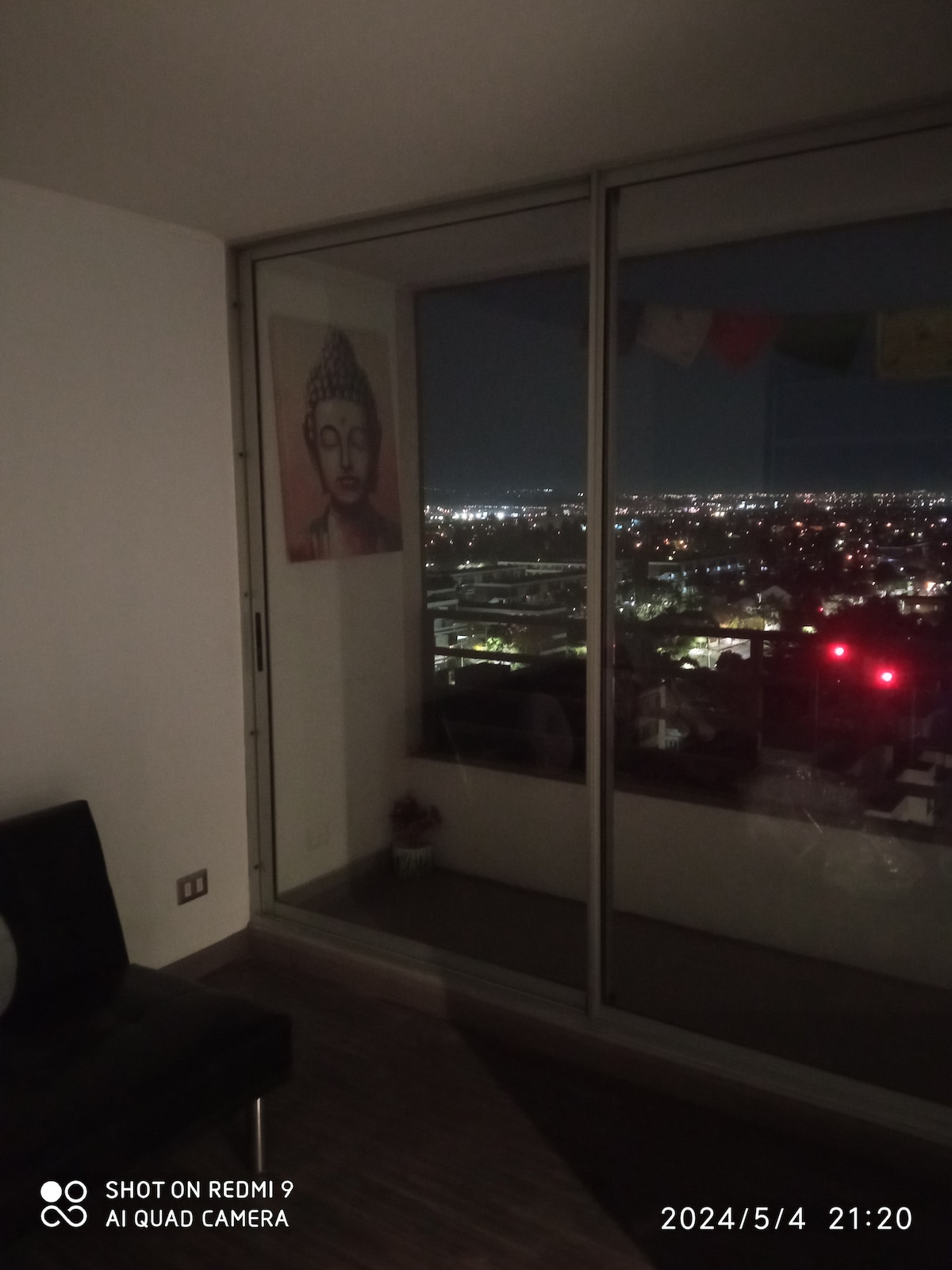 A sleek living area is visible, featuring large glass windows that offer a panoramic view of the city lights at night. A piece of art depicting a serene figure hangs on the wall, complementing the modern decor of the room.