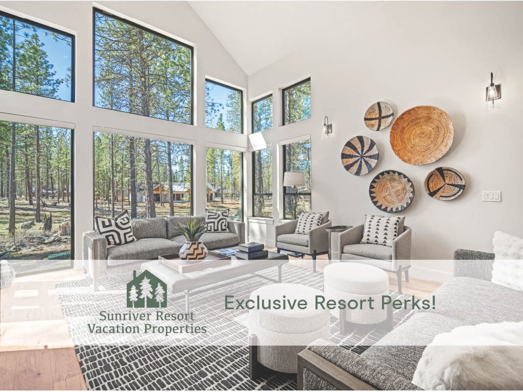 A bright living area is surrounded by large windows that showcase the wooded landscape. Modern furnishings include a sectional sofa, accent chairs, and a coffee table, while decorative wall art adds contrast to the neutral palette. Natural light enhances the inviting atmosphere.