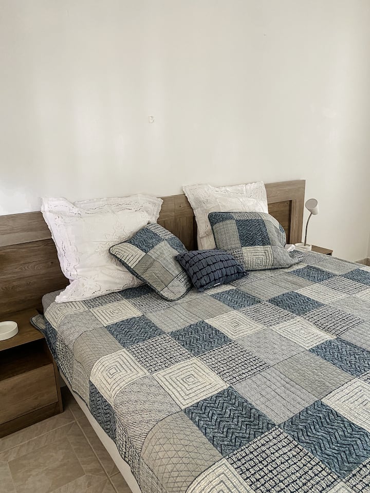 Ova mirna spavaća soba ima veliki krevet s plavim i sivim poplunom i raznim jastucima postavljenim na drvenom uzglavlju. Jednostavan noćni ormarić s modernom svjetiljkom nadopunjuje čiste, bijele zidove i svijetle pločice, stvarajući miran prostor.