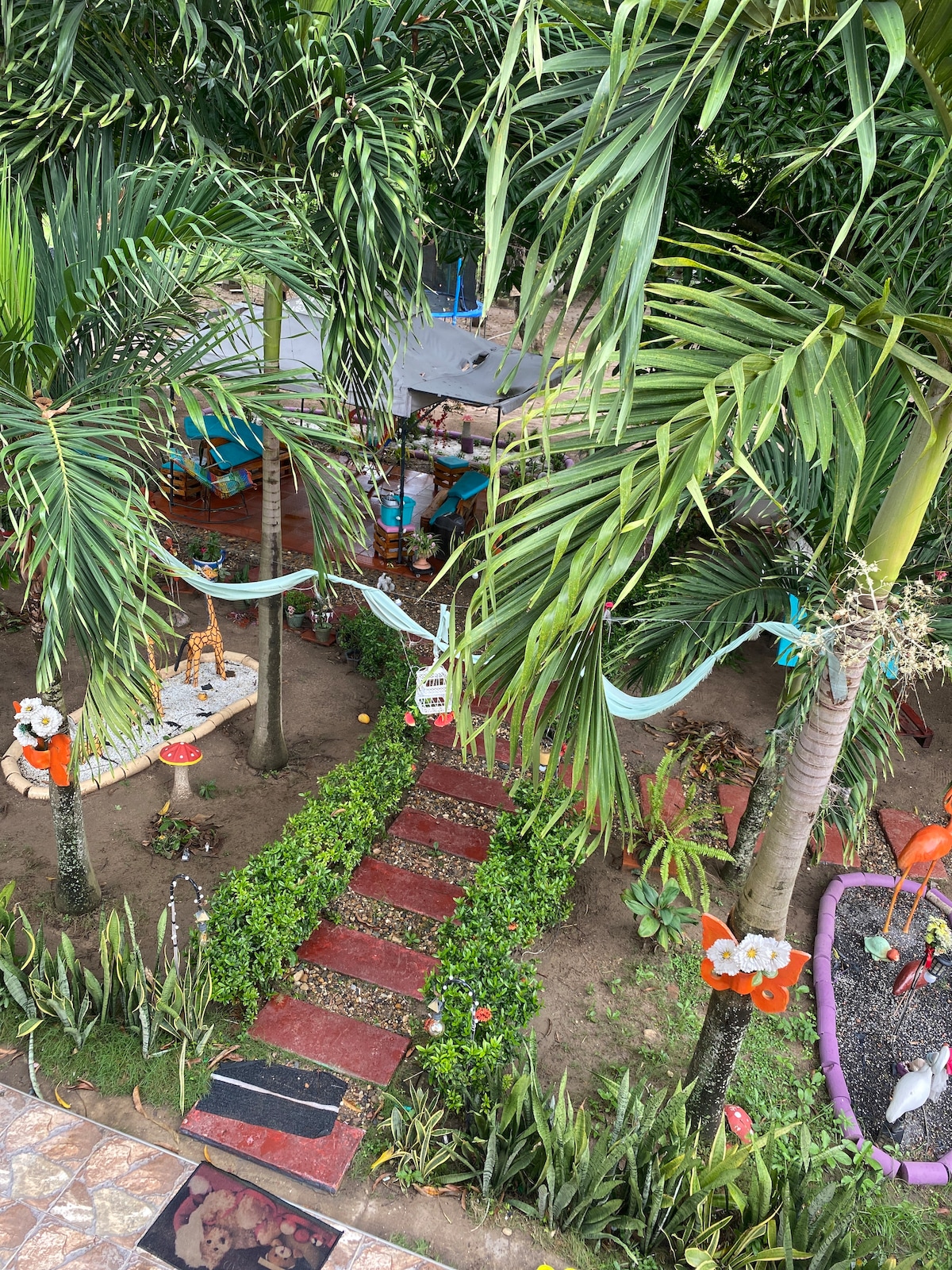 A lush garden features well-maintained plants and vibrant decorative elements. A pathway lined with greenery leads to a shaded seating area surrounded by tall palm trees. Colorful decorations, including flowers and whimsical figures, enhance the natural beauty of the outdoor space.