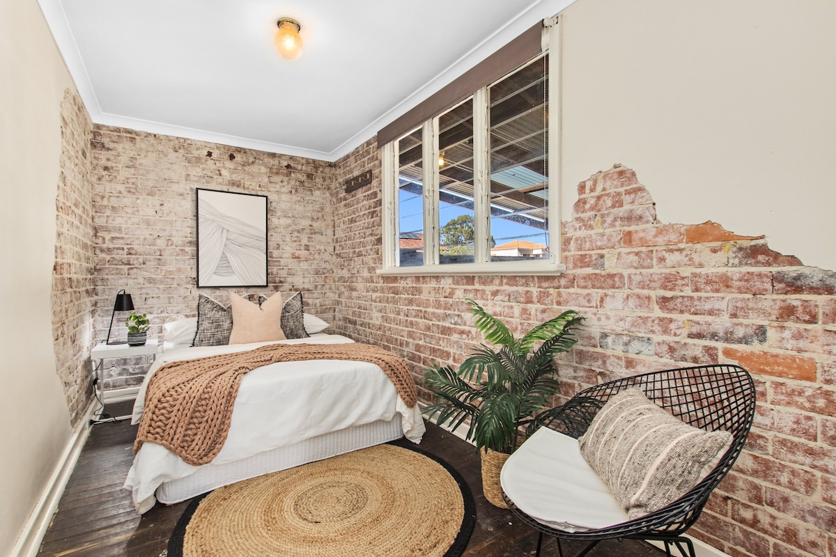 A cozy bedroom features exposed brick walls, adding character to the space. A spacious bed is dressed with neutral-toned linens, complemented by a textured blanket. A round jute rug anchors the room, while a chair and potted plant contribute to its inviting atmosphere.