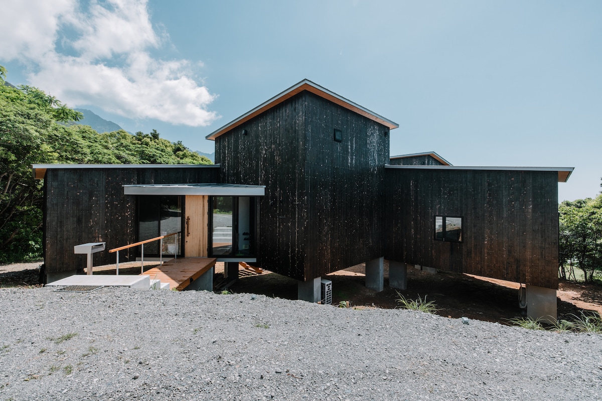 A modern structure features a striking black exterior with wooden accents. The roof showcases unique angles, while large glass doors provide a view into the open interior. A gravel path leads to the entrance, surrounded by lush greenery and a clear blue sky.