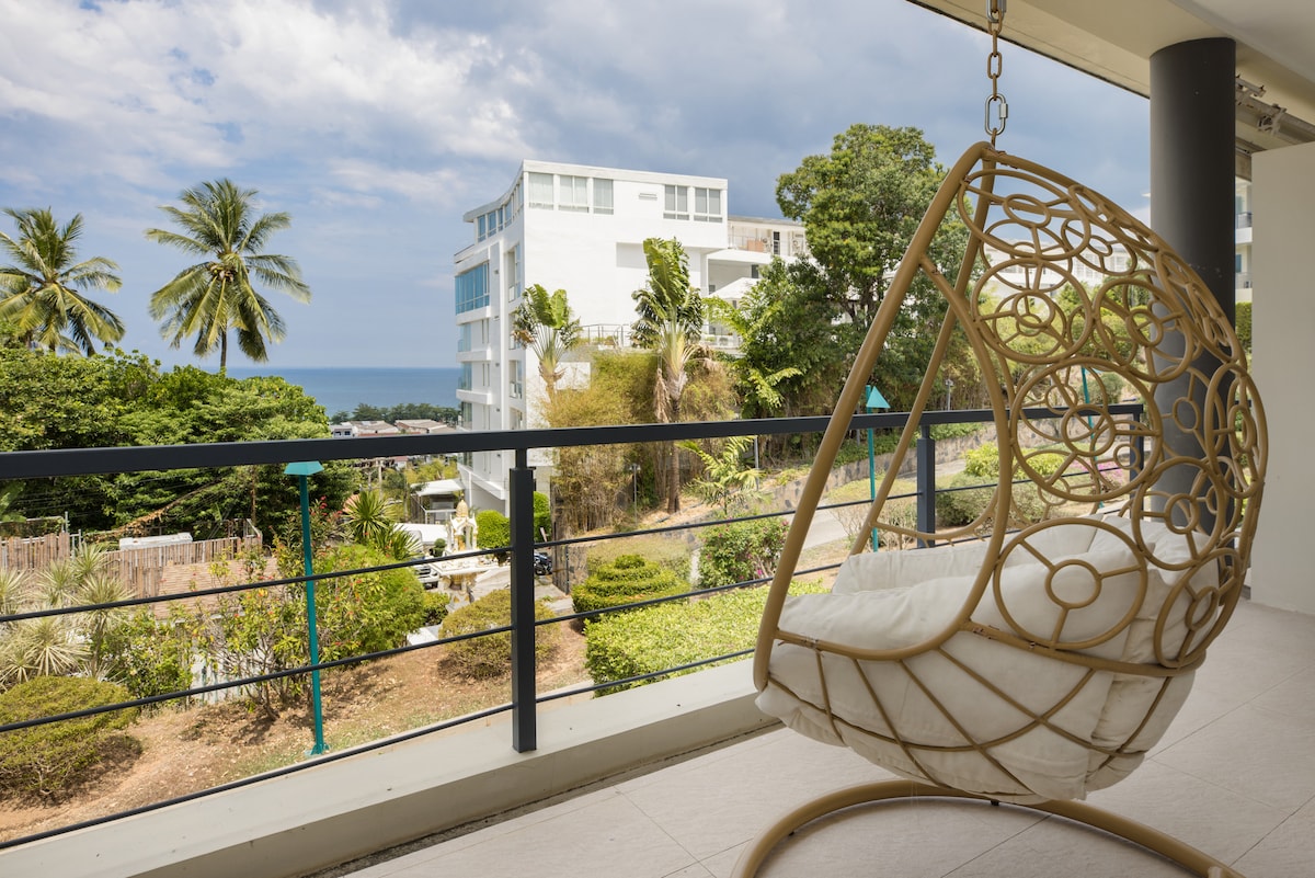 A stylish swing chair is positioned on the balcony, offering views of lush greenery and the ocean beyond. The surrounding area features palm trees and manicured landscaping, creating a serene backdrop for relaxation.