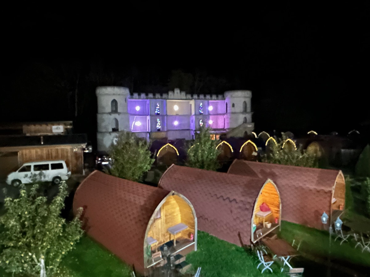 Cozy wooden barrel-huts are arranged in a grassy area, illuminated softly at night. In the background, a castle-like structure features colorful lighting, enhancing the whimsical atmosphere. Outdoor seating is present near the huts, presenting a welcoming space for relaxation and gathering.