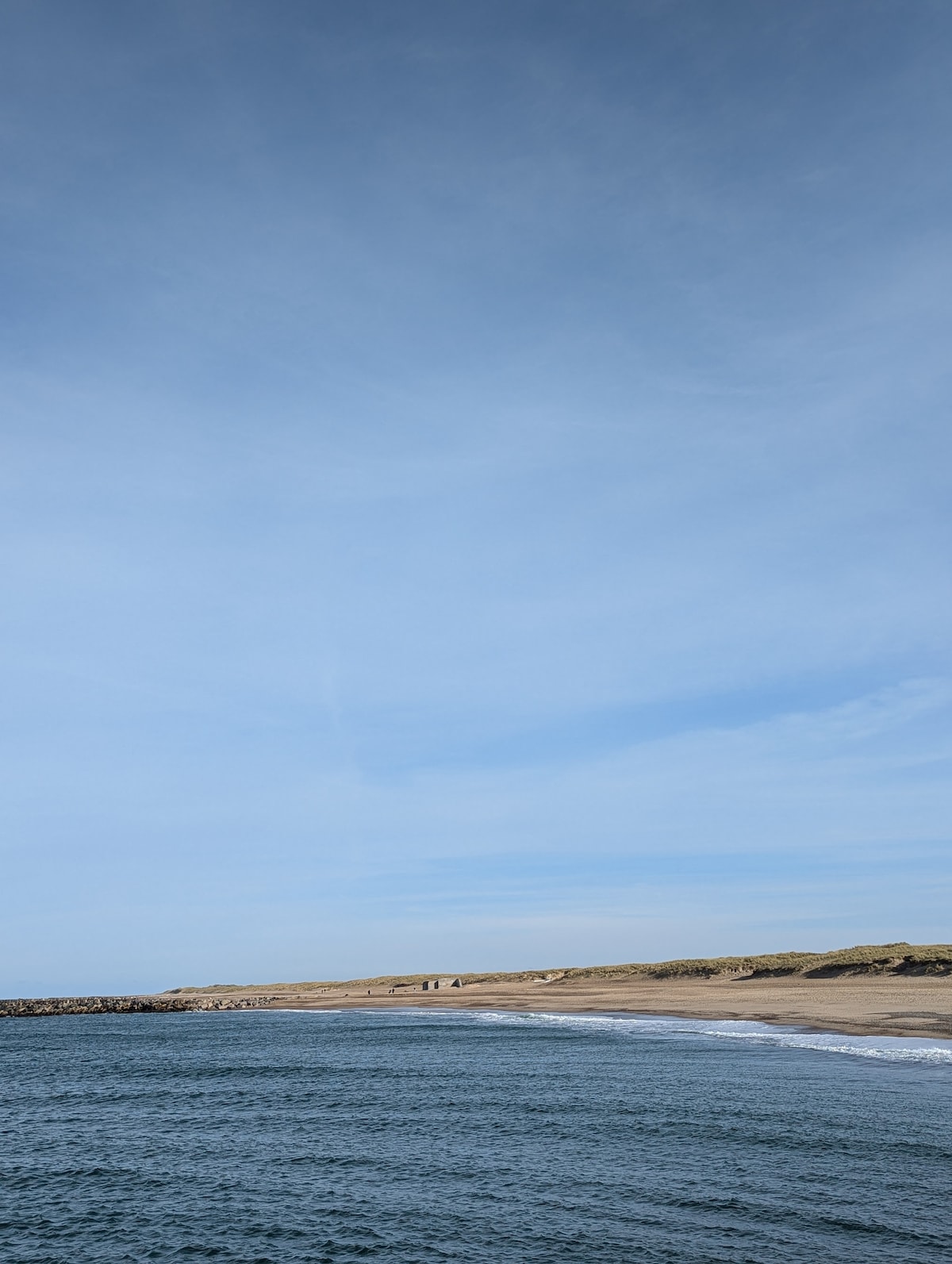 A serene coastal view is presented, featuring calm blue waters meeting a sandy beach under a clear sky. Gentle waves lap at the shore, while distant sand dunes are visible along the horizon. The overall atmosphere reflects a tranquil shoreline.
