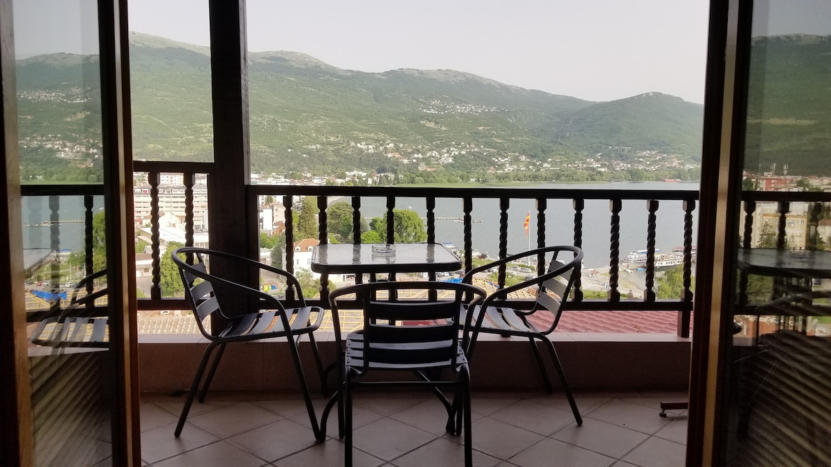 A balcony features a glass table and two chairs, offering a view of the lake and surrounding mountains. The area is shaded by wooden railings, and the landscape showcases lush greenery and distant hills.