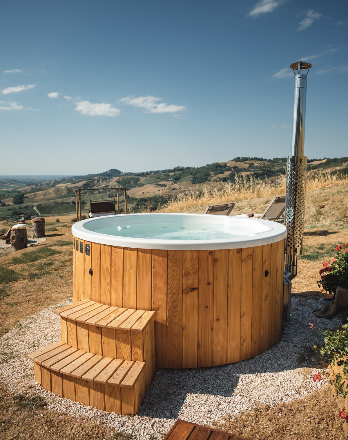 A wooden hot tub is positioned outdoors, surrounded by rolling hills and open sky. Steps lead up to the tub's edge, and lounge chairs are placed nearby, providing a serene setting for relaxation amidst nature.