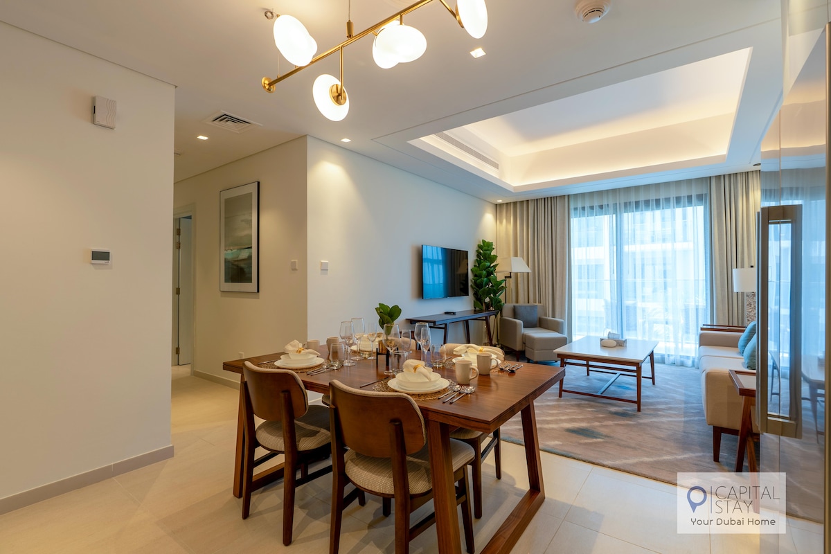 A modern dining area is set with a wooden table that accommodates six chairs. Soft natural light fills the space through large windows, while a comfortable seating area with a neutral palette is visible in the background.