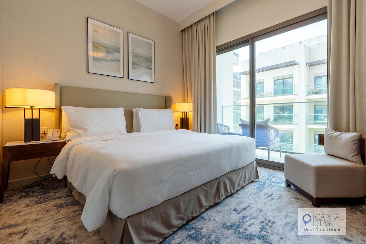 A well-appointed bedroom features a spacious bed with white linens and two bedside lamps providing soft illumination. Large windows allow natural light to fill the room, with views onto a balcony furnished with two chairs. Neutral tones create a calming environment.