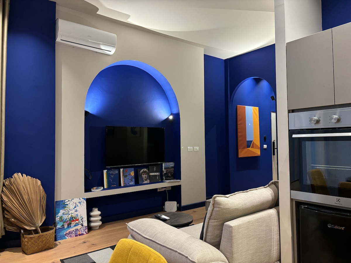A modern living room area features deep blue walls paired with soft accent lighting. A sleek TV is mounted on the wall above a minimalist shelf displaying colorful art books. A comfortable sofa invites relaxation, while stylish decor elements add artistic flair to the space.