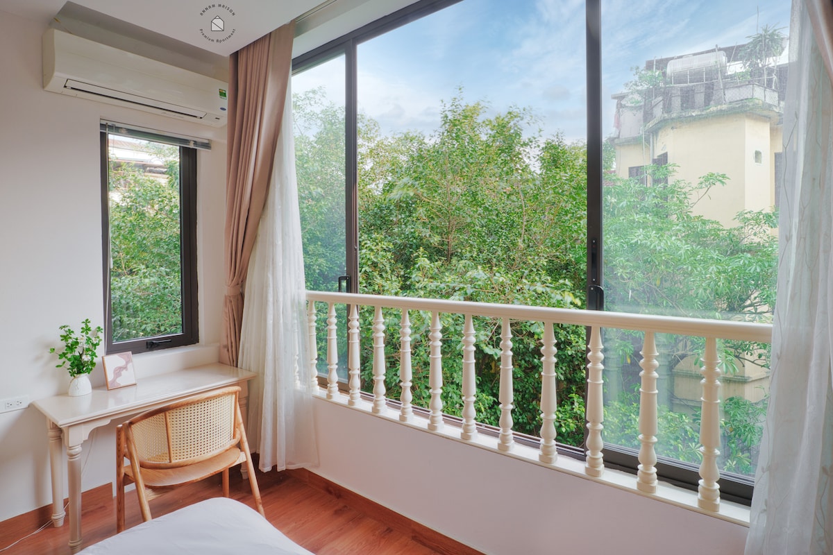 Large windows frame views of lush greenery outside the room. A modest desk is positioned near the window, while soft curtains gently drape along the sides. Natural light brightens the space, highlighting the wooden flooring and minimalist decor.