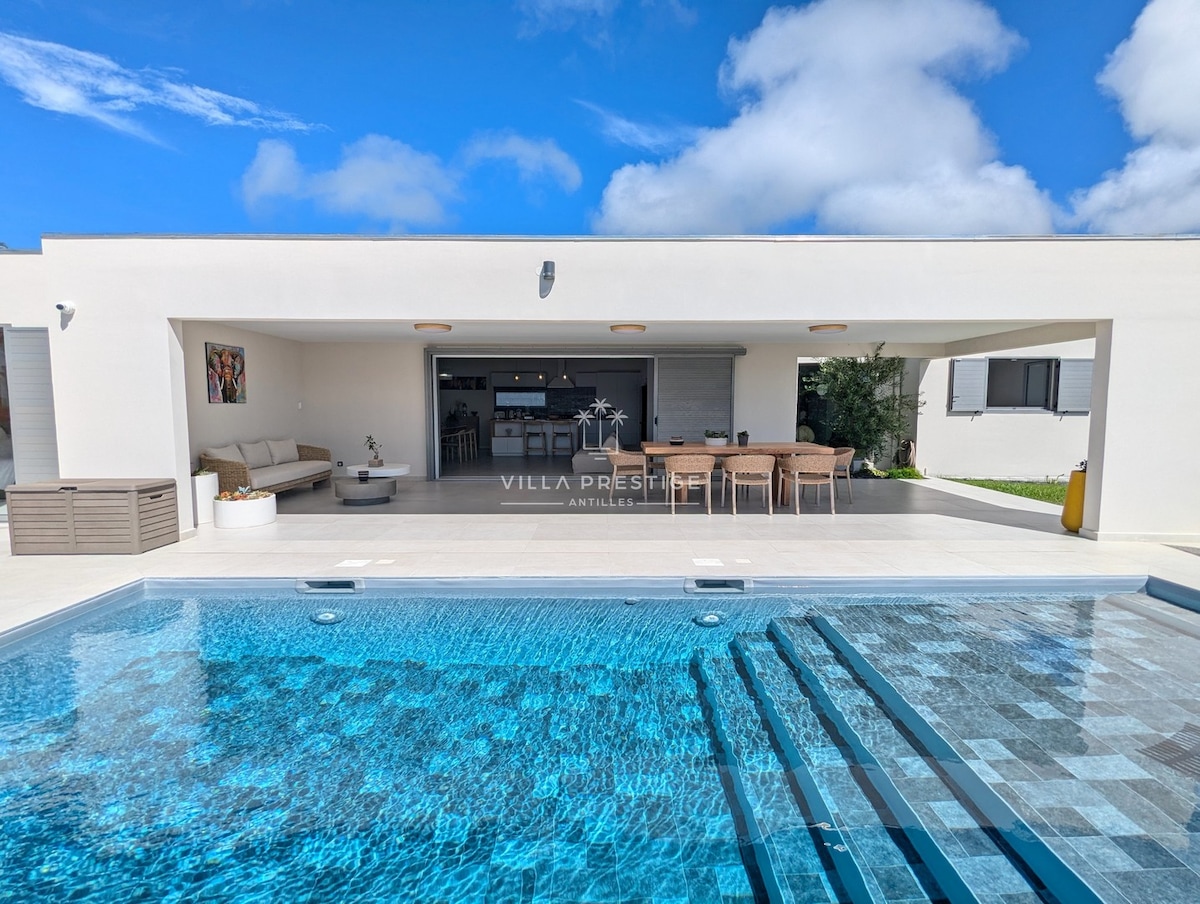 A clear view of a modern villa's outdoor area is displayed, featuring a shimmering pool with steps leading in. A covered terrace adjacent to the pool includes a comfortable seating area and a dining table, all set against a backdrop of blue skies and soft clouds.