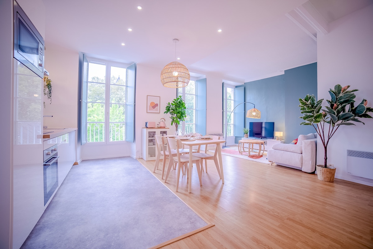 A bright and spacious living area is visible, characterized by large windows that invite natural light. A dining table is arranged for four, alongside a comfortable sofa. Stylish decor includes a leafy plant and modern light fixtures, creating a welcoming space for relaxation.