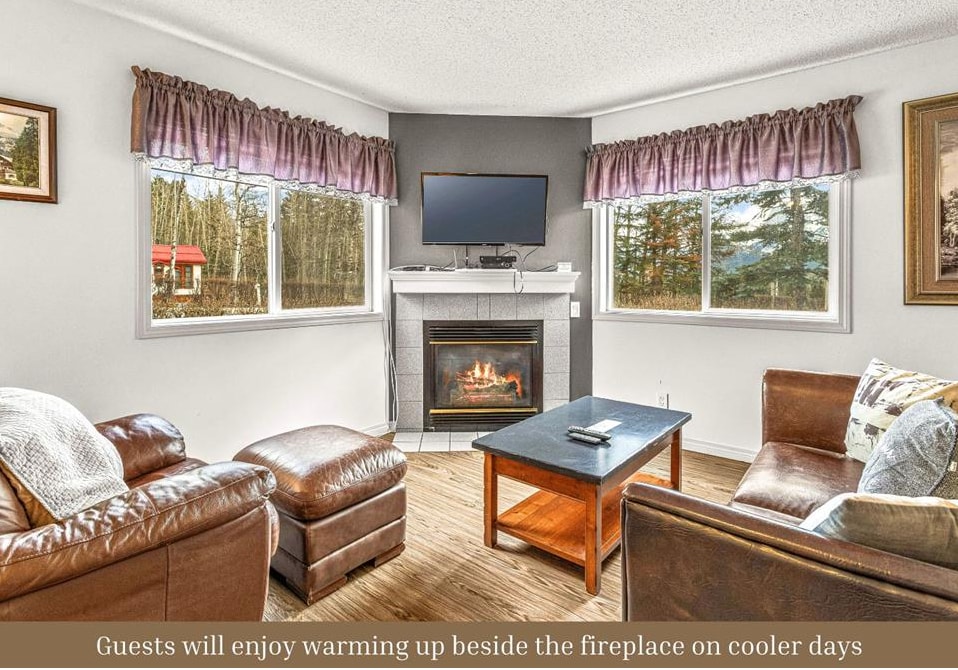 A comfortable living area features a cozy fireplace, with a mounted TV above it. Large windows provide views of the surrounding greenery. A coffee table sits between two leather sofas, inviting relaxation and conversation.