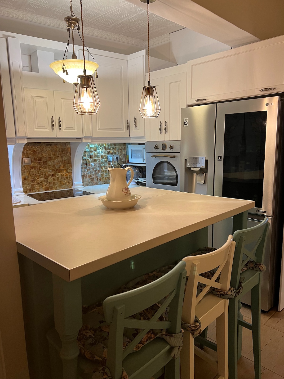 A well-equipped kitchen is presented, featuring a light wooden island with three chairs. The cabinets are white, and pendant lights hang above. A stainless steel refrigerator stands beside a built-in oven, while warm-toned backsplash tiles add a subtle glow to the area.