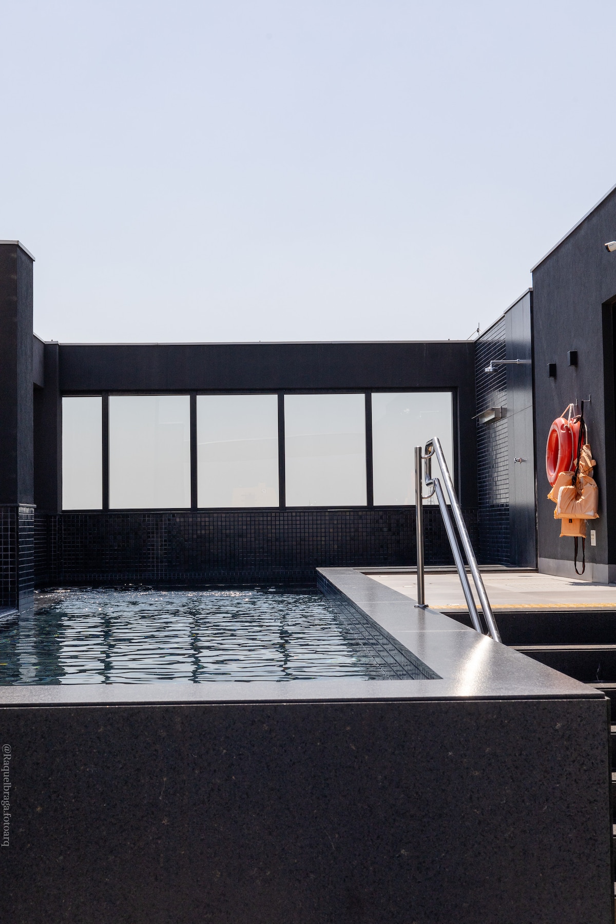 A sleek rooftop pool is framed by modern architecture. Clear water reflects the sunlight, and views are framed by large windows. Safety equipment, including life rings, is visible on the wall, enhancing accessibility. The overall design promotes a serene and inviting environment.