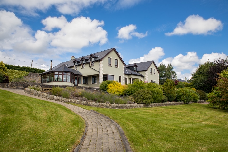 A spacious home is set within lush gardens, featuring a winding stone path leading to the entrance. The structure showcases a mix of contemporary and traditional architectural styles with ample windows, allowing natural light to fill the living spaces.