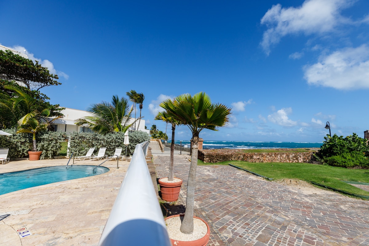 A serene outdoor area is observed, featuring a swimming pool surrounded by lounge chairs. Lush greenery is present alongside a palm tree. The ocean is visible in the background, under a clear blue sky with scattered clouds.