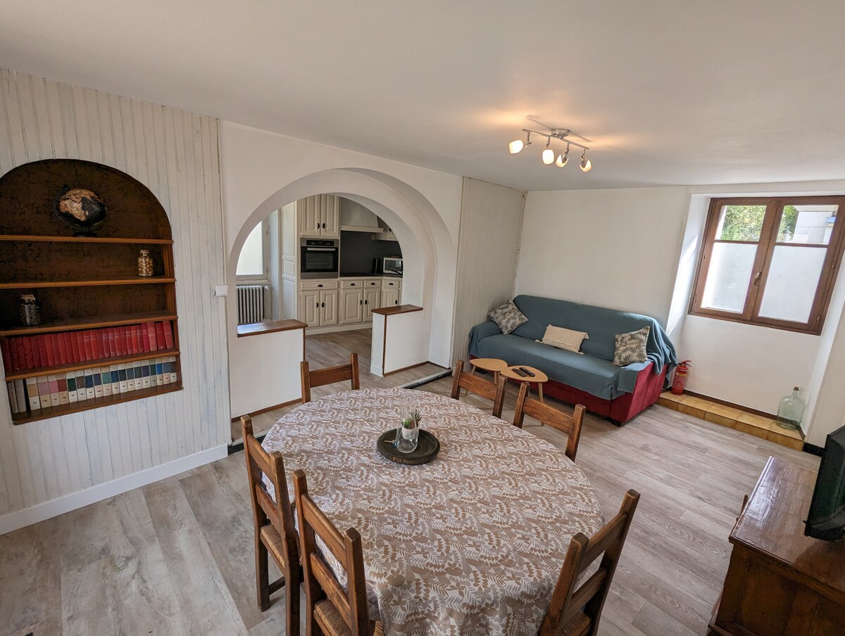 A spacious dining area is featured, showcasing a round table surrounded by wooden chairs. Nearby, a comfortable sofa adds to the living space, while an archway leads to the kitchen. Natural light filters through windows set in the walls, enhancing the atmosphere.