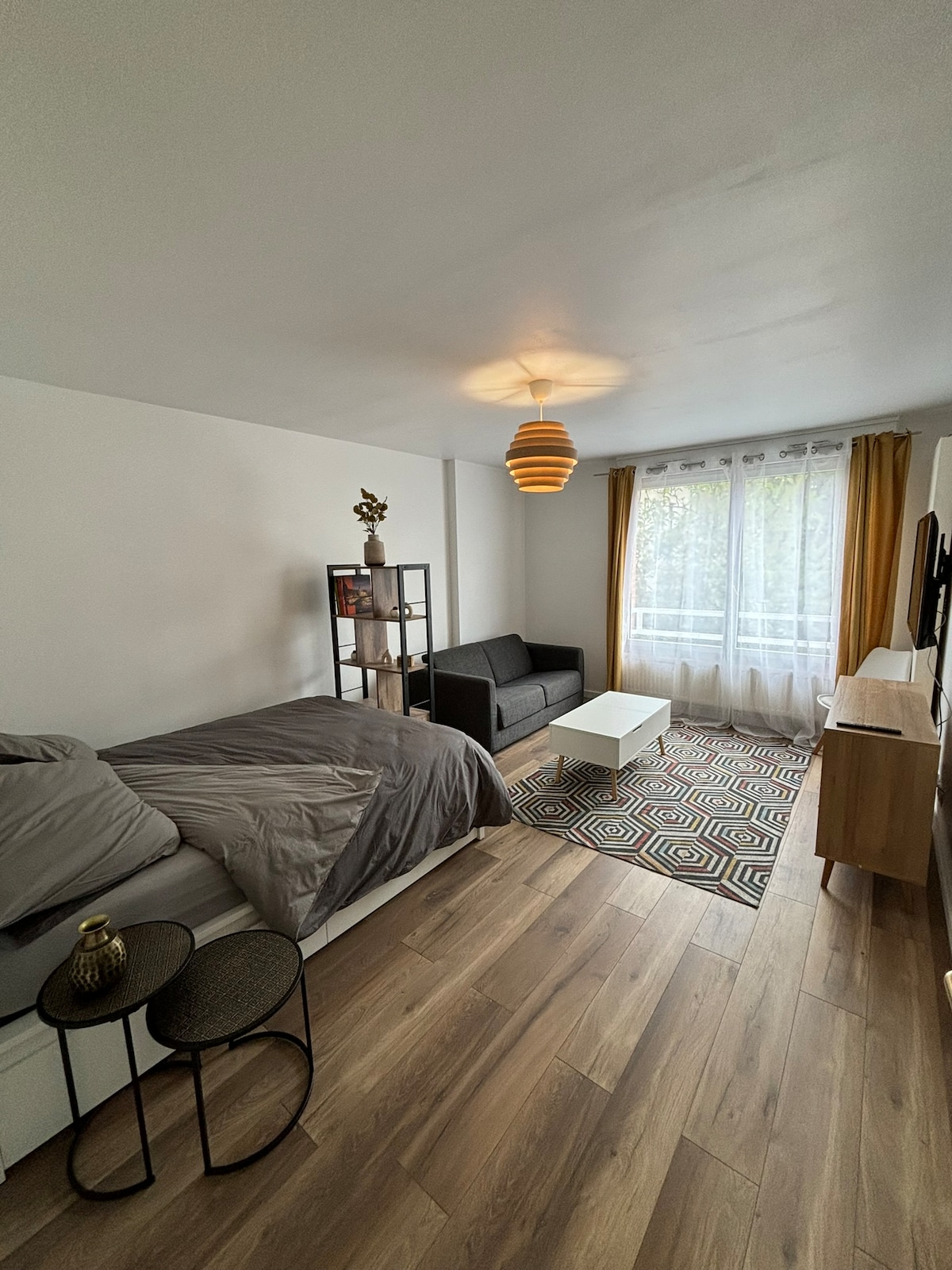 A modern studio space features a spacious bed with neutral bedding, accompanied by a sleek black sofa. A light wood coffee table sits on a patterned rug, while a decorative shelf displays a small statue. Natural light filters through large windows, framed by yellow curtains.