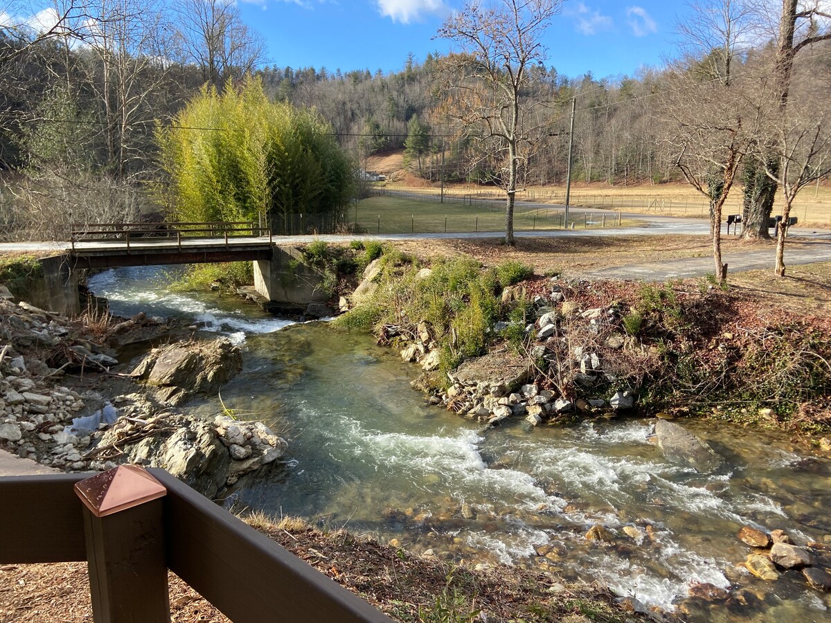 A serene stream flows gently through a natural landscape, flanked by rocks and sparse vegetation. A wooden bridge arches over the water, leading to an open field surrounded by trees. Clear blue skies provide a bright backdrop to the tranquil setting.