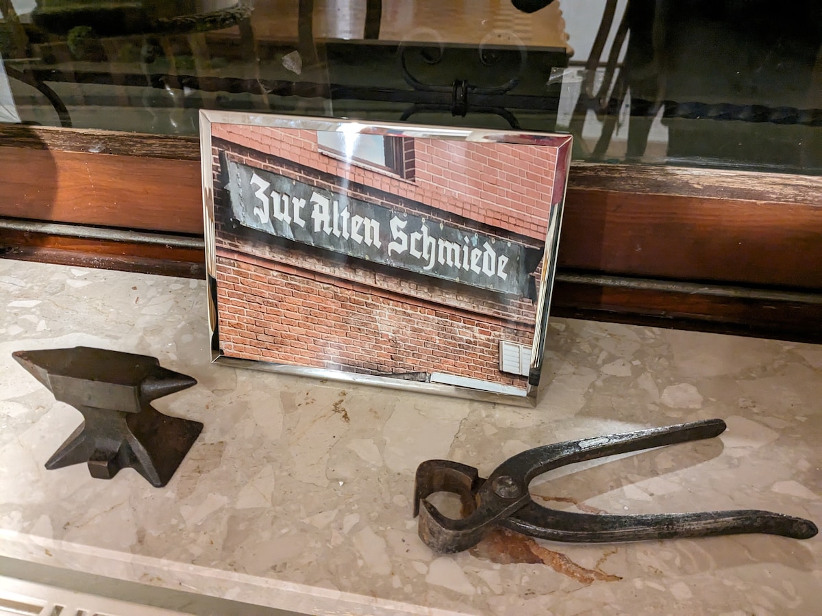 A photograph featuring the rustic sign 'Zur Alten Schmiede' is displayed prominently in a frame. Next to it, a small anvil and a pair of vintage tongs showcase the historical craftsmanship associated with the house.