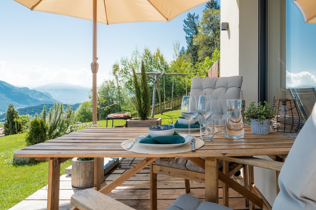 A wooden table is set on a terrace, accompanied by comfortable seating. Glassware and dishware are arranged invitingly. The surrounding garden showcases greenery and scenic views of the valley, with an umbrella providing shade from the sun.