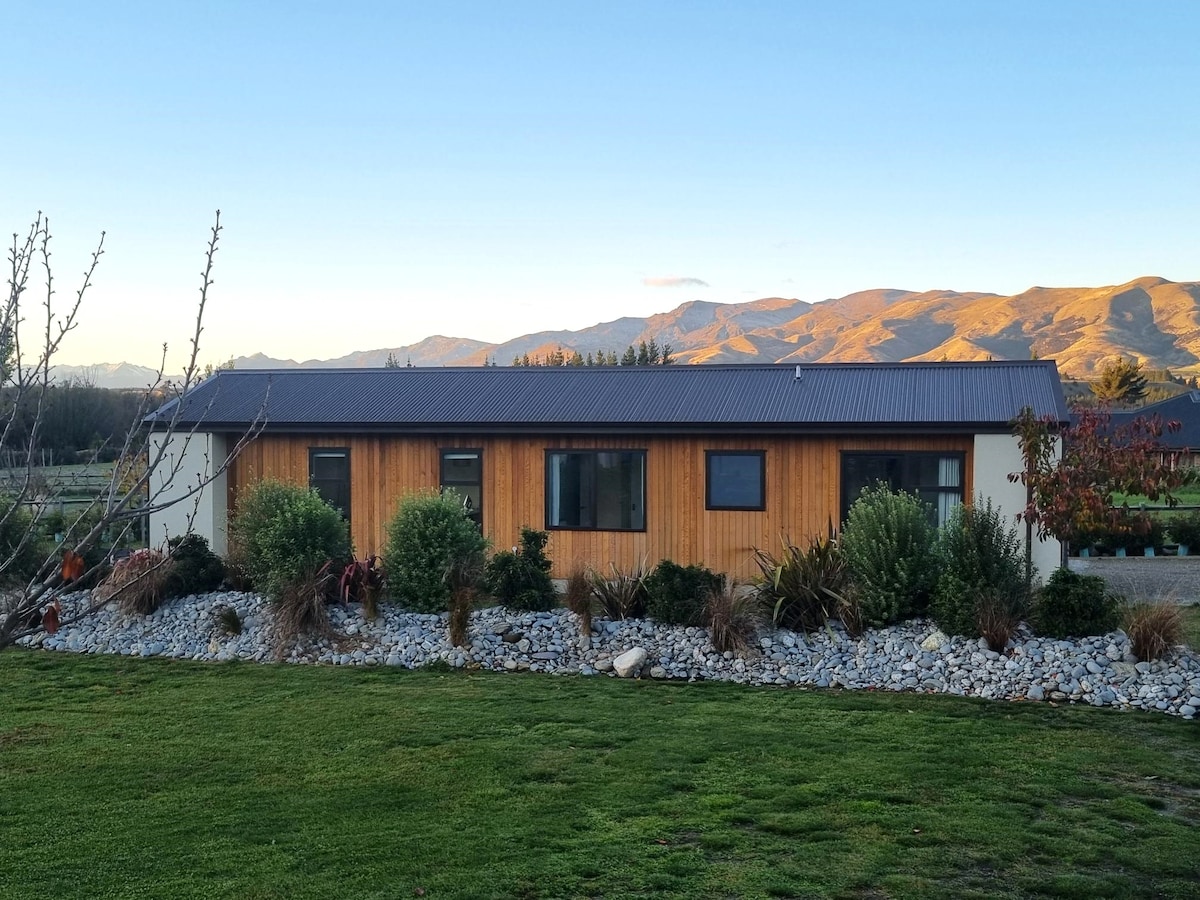 The exterior of a modern Scandinavian-style house is showcased, featuring a wooden facade and a dark roof. Surrounding the house are landscaped gardens with various shrubs and rocks, all set against a stunning backdrop of rolling hills and mountains under a clear sky.
