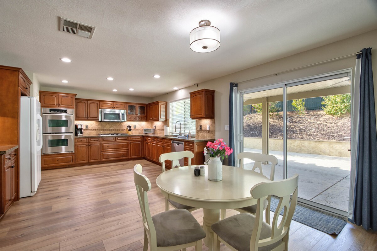 An open kitchen features rich wood cabinetry and stainless steel appliances. A round dining table with four chairs is positioned to the side, complemented by a vibrant vase of flowers. Natural light streams through sliding glass doors leading to the patio.