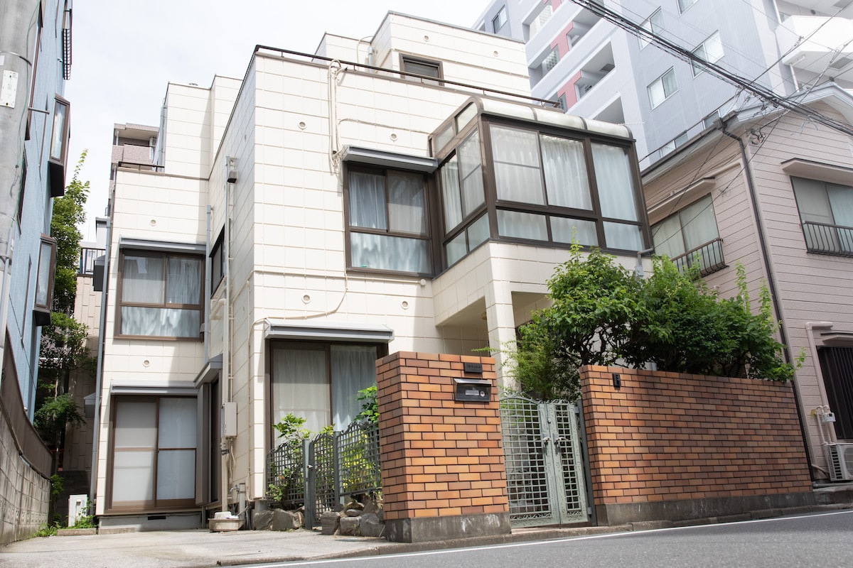 The exterior of a three-story building is shown, featuring a blend of light-colored walls and large windows. A brick wall with a gate surrounds the entrance, while greenery is visible near the base of the structure. The surrounding area includes neighboring buildings.