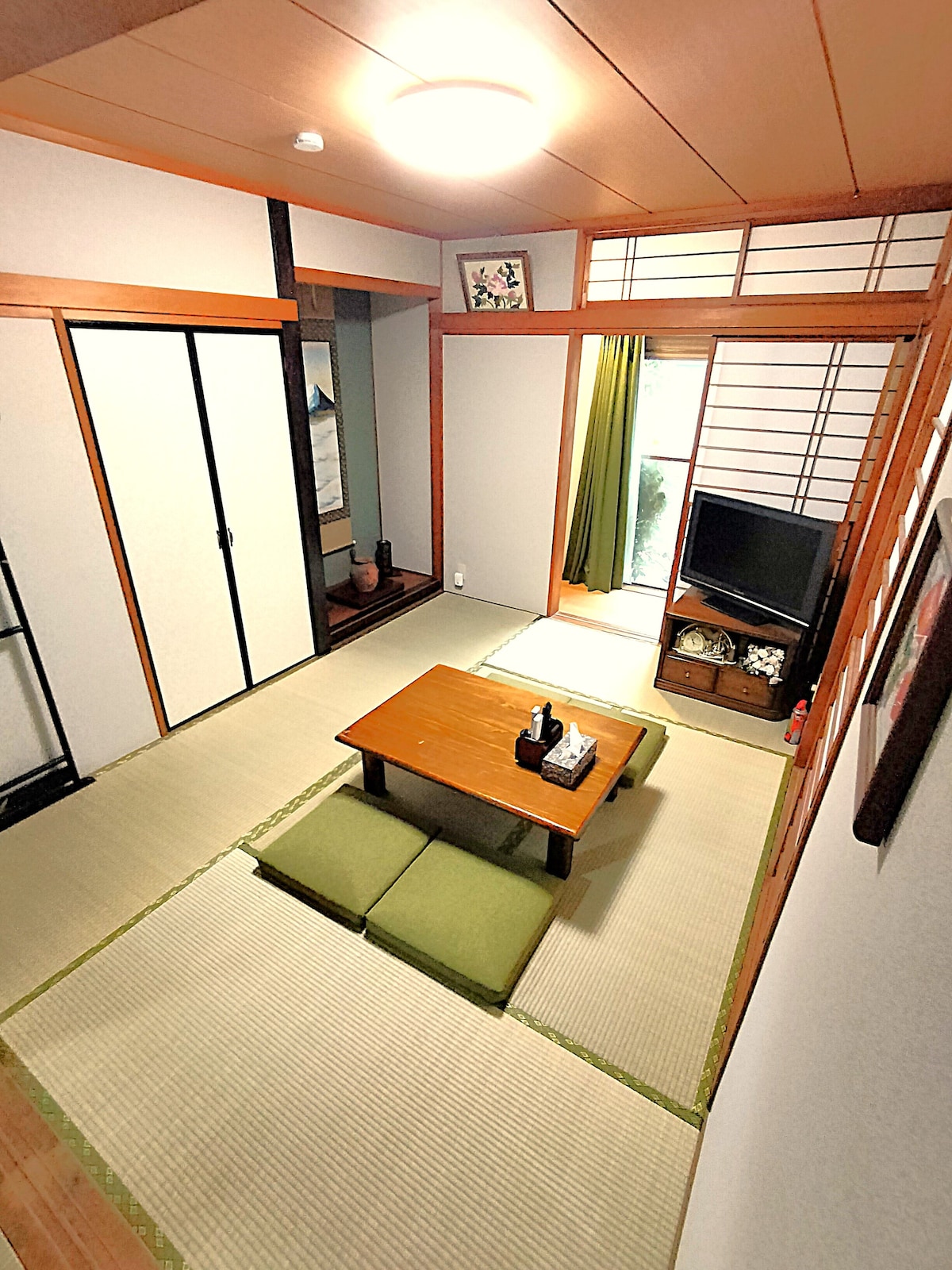 A traditional Japanese room features tatami flooring and sliding doors, complemented by natural light from a window with green curtains. A low wooden table is set atop the tatami, surrounded by floor cushions. A television and minimalist decor add to the cozy ambiance.