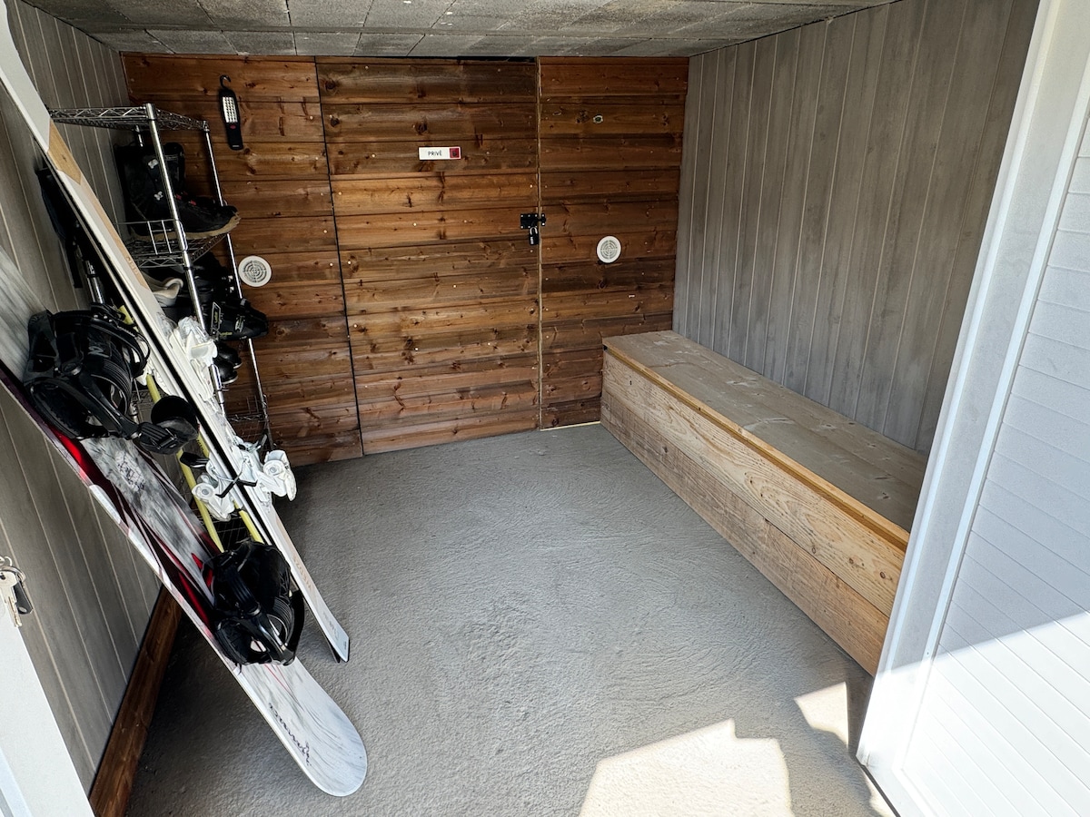 A ski storage area is visible, featuring wooden walls and benches. Snowboards are positioned against the wall, while a bench provides seating for equipment storage. Shelving units are included for additional gear storage. The entrance is secured with a lock for safety.
