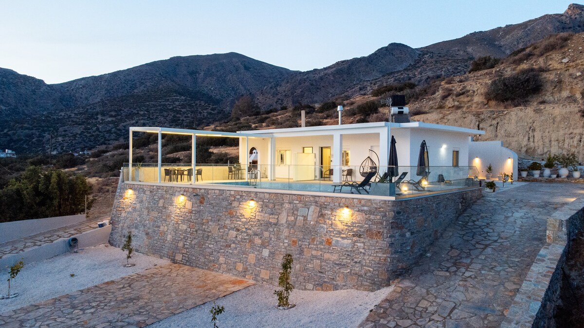 The image captures a modern villa nestled against a mountainous backdrop, showcasing a spacious terrace with seating. Soft lighting highlights the stone walls, while a stone pathway leads up to the inviting entrance. The surrounding area features landscaped gardens, enhancing the tranquil atmosphere.