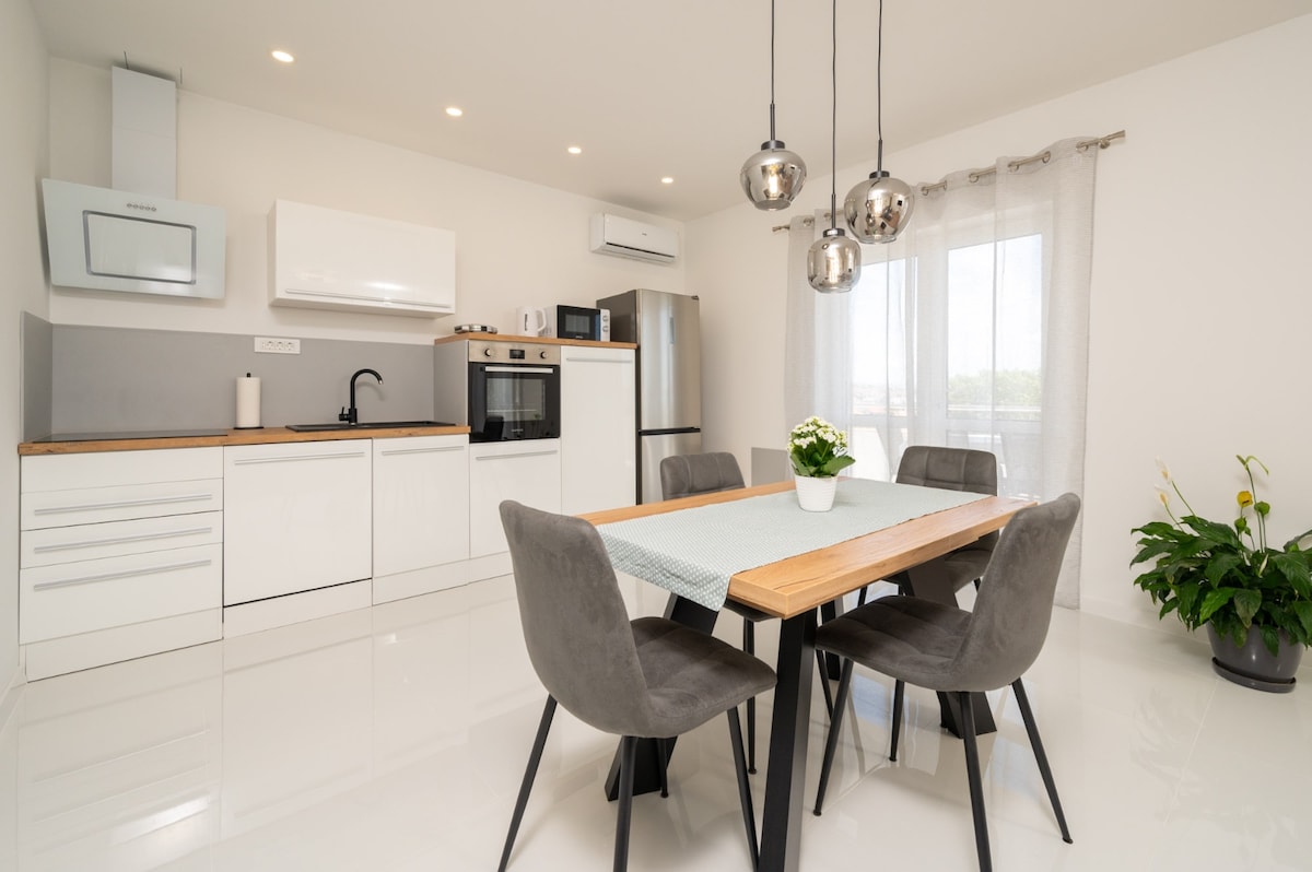 A modern kitchen and dining area features a sleek white cabinetry setup, complemented by a wooden countertop. A dining table with four chairs is positioned beneath stylish pendant lights. Natural light fills the space through a nearby window, accentuating the minimalist design.