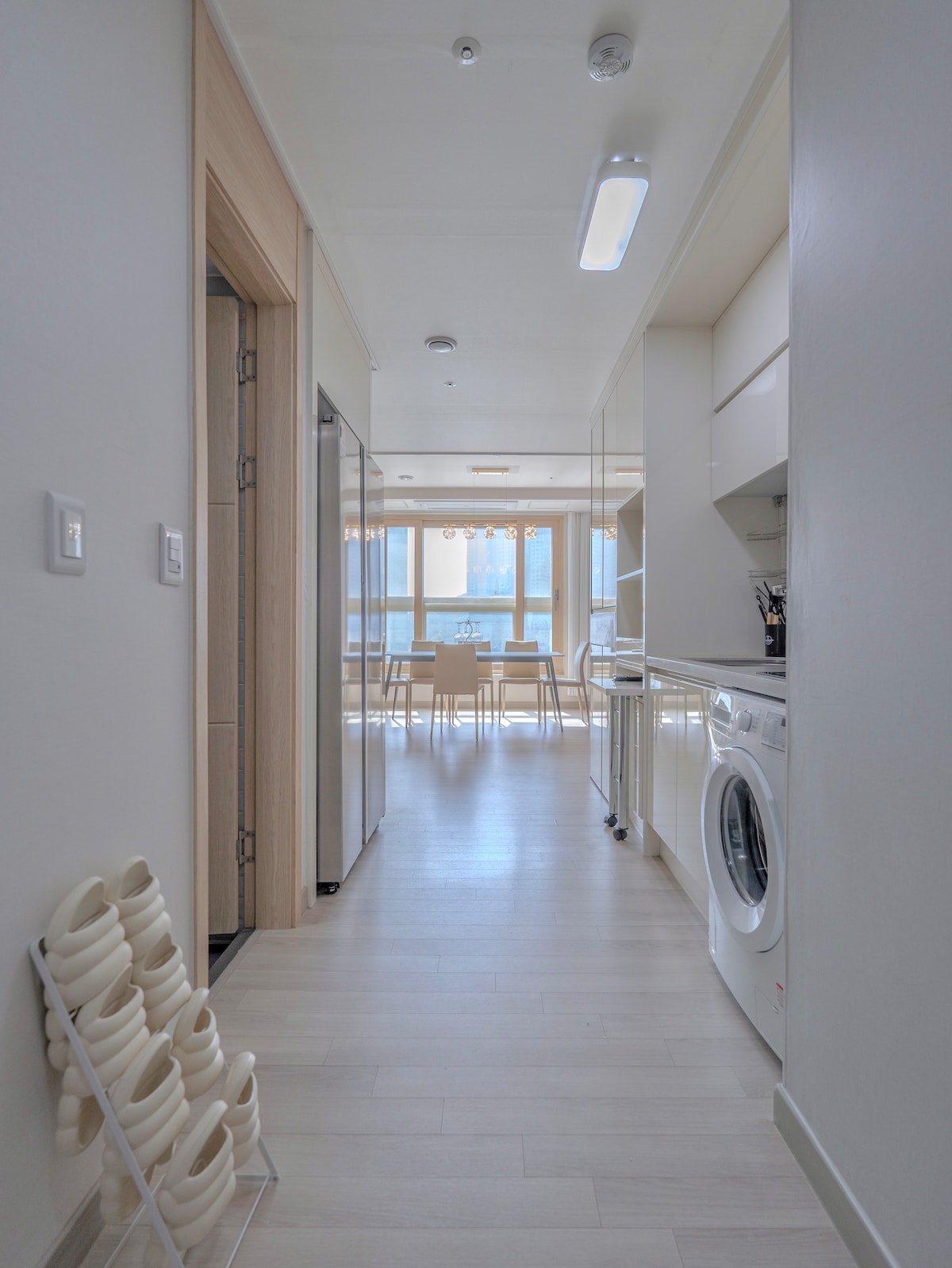 A bright hallway features a modern design with a sleek washing machine and white cabinetry. Light-colored flooring enhances the spacious feel. At the end of the hallway, a dining area is visible, showcasing a table surrounded by chairs and illuminated by natural light through large windows.