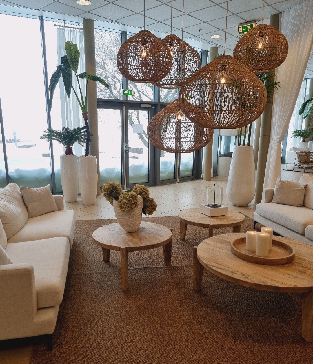 A modern lounge area features light-colored sofas arranged around round wooden tables. Large woven pendant lights hang above, offering a natural touch. Elegant plants are displayed in tall white vases, while large windows provide a view of the snowy outdoors.