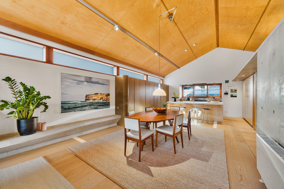 A bright dining area features a round wooden table surrounded by four chairs, with a large indoor plant adding a touch of greenery. Natural light floods in through multiple windows along the ceiling, illuminating the polished wooden surfaces and soft textures of the area rug.