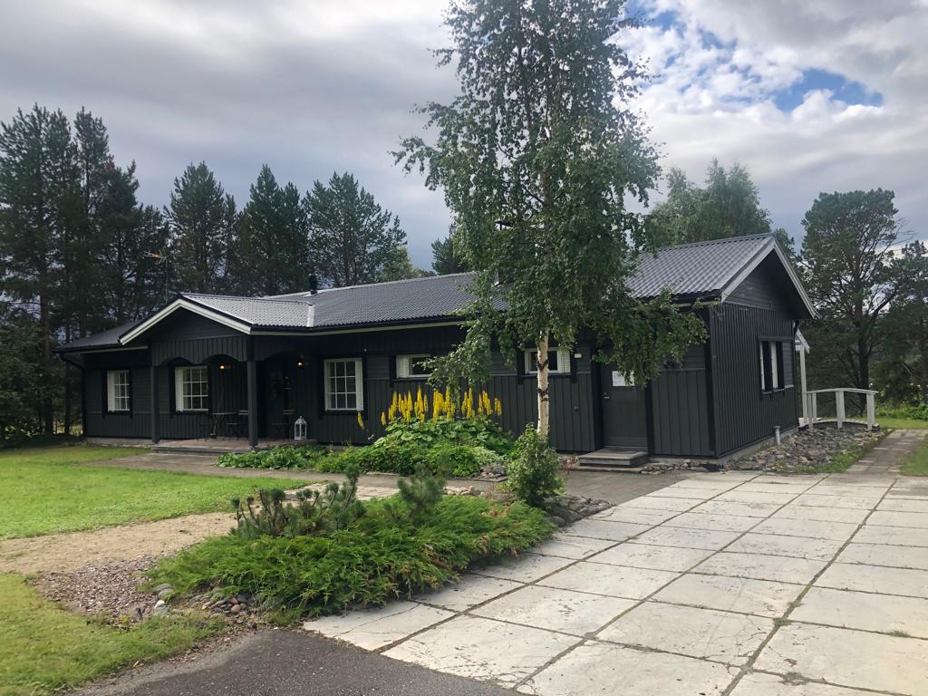 A charming villa with dark wood siding is set amidst lush greenery and tall trees. The front yard features carefully tended flower beds and a wide driveway leading up to the entrance, creating a welcoming approach to the home.