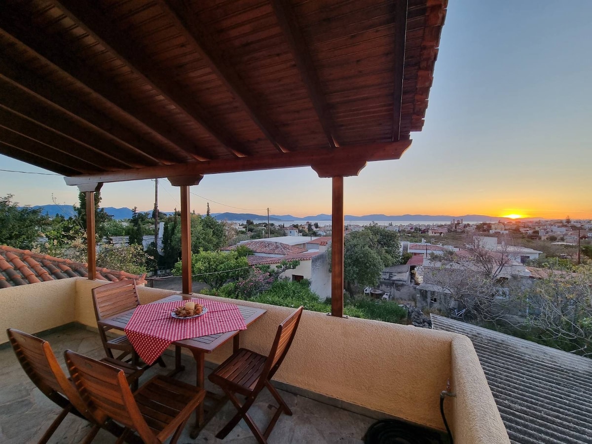 A spacious terrace is shown, furnished with a round table and wooden chairs, offering a scenic view of the landscape. The sun sets in the background, casting warm hues across the sky while the rooftops of nearby buildings are partially visible.