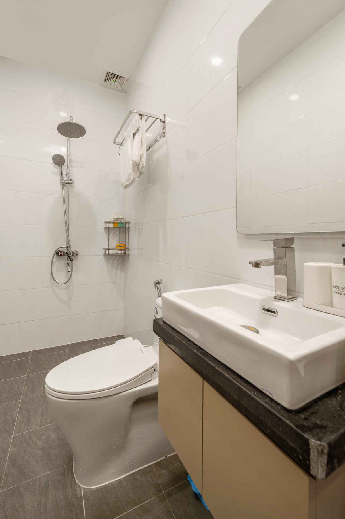 A modern bathroom design features a walk-in shower with a sleek rain showerhead and clear glass partition. A white basin is placed on a dark countertop, accompanied by neatly arranged toiletries. Bright lighting highlights the clean tile walls and floor.