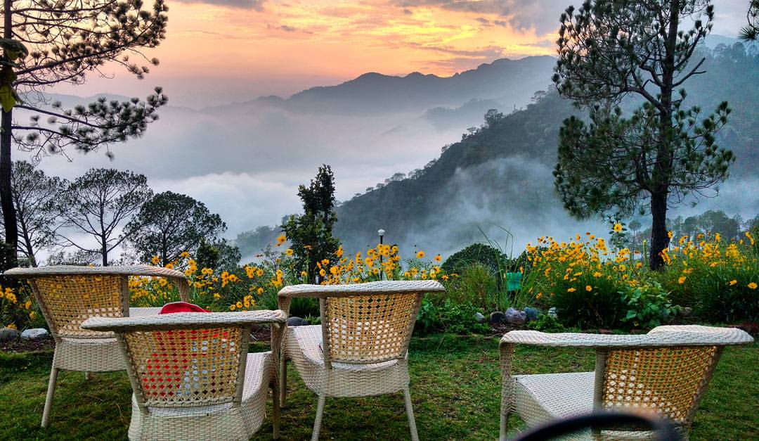 A serene outdoor seating area features four woven chairs facing a view of rolling hills and a colorful sunset. Vibrant flowers bloom in the foreground, while mist hangs softly over the landscape, creating a peaceful, natural setting.