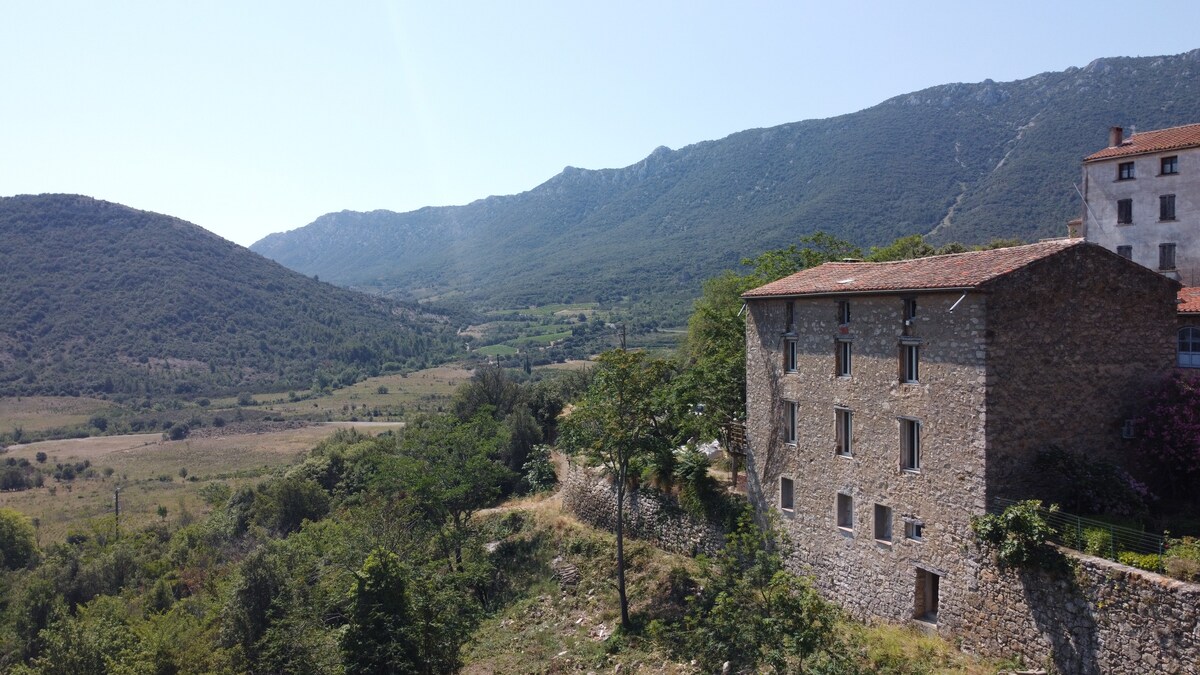 A rustic stone house is positioned on a gentle slope, surrounded by greenery and framed by distant mountains. The landscape showcases a blend of hills and valleys, with a clear blue sky above, creating a serene outdoor environment.