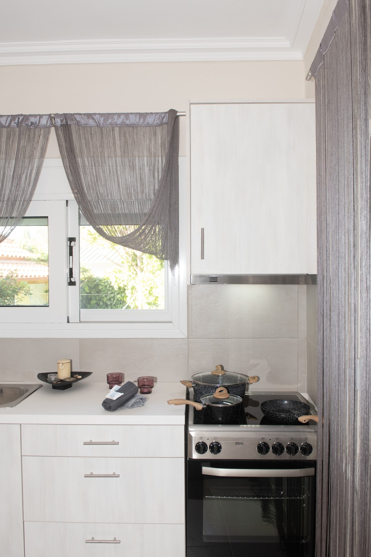 The kitchen features modern cabinetry with sleek white finishes, complemented by a light grey backsplash. A window allows natural light to brighten the space, adorned with sheer curtains. Cooking essentials, including pots and utensils, are organized on the countertop, enhancing practicality.