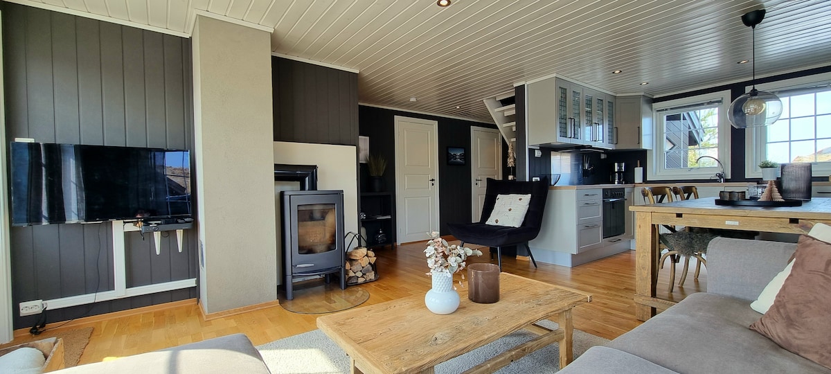 An open living area features a comfortable seating arrangement and a wooden coffee table. A modern kitchen is visible in the background, along with a cozy wood-burning stove. Large windows allow natural light to fill the space, highlighting the wood flooring and neutral color scheme.