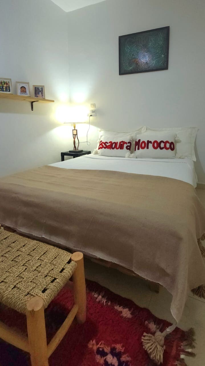 A cozy sleeping area is presented with a spacious bed covered by a light blanket. Decorative pillows with 'Essaouira Morocco' are placed against the headboard. A small woven chair is positioned beside the bed, and a shelf with framed photos is mounted on the wall.