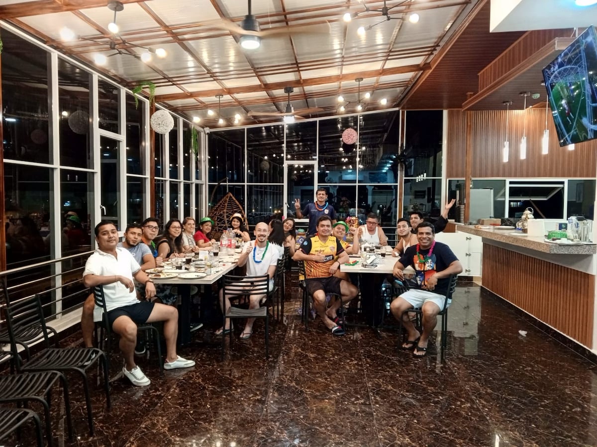 A spacious dining area is presented, featuring a long table set with plates and utensils. Large windows allow natural light to illuminate the space. Guests are seated around the table, enjoying a meal in a modern environment with wood accents and a sleek countertop in the background.